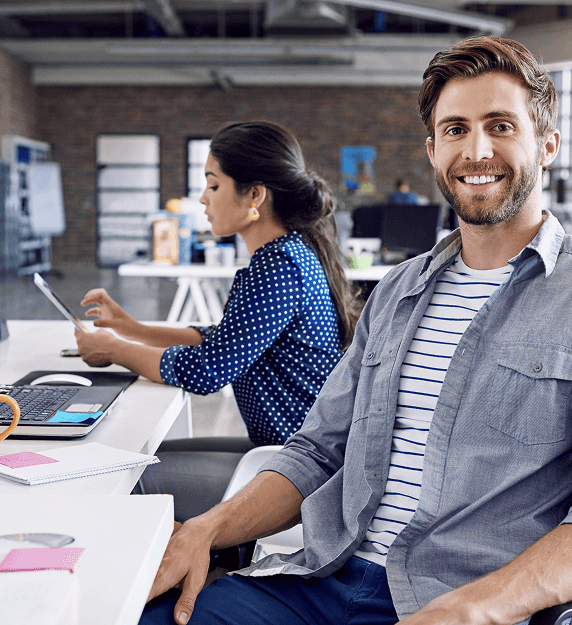 dos jóvenes profesionales trabajando en una oficina de estilo industrial; un hombre sonríe a la cámara mientras una mujer utiliza una tablet al fondo