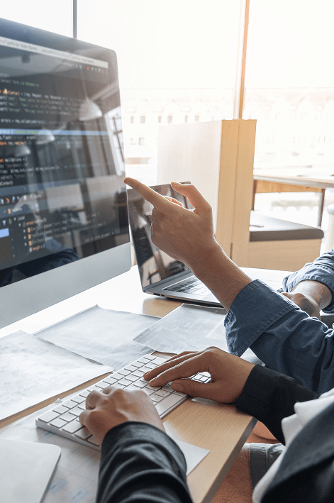 primer plano de dos personas colaborando frente a un monitor que muestra código de programación; una persona teclea mientras la otra señala la pantalla en una oficina iluminada