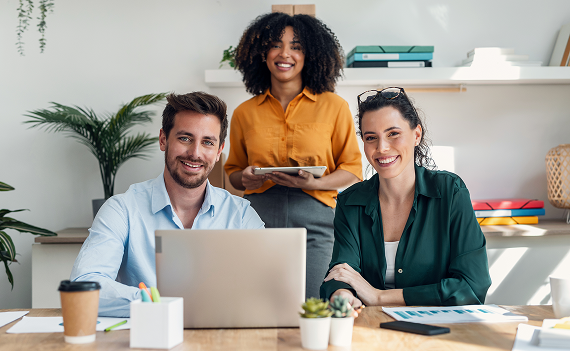tres profesionales sonrientes posando en una oficina luminosa con una computadora portátil y una tablet